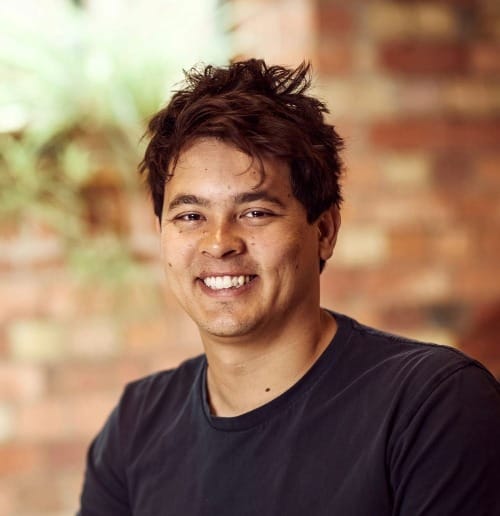 Man with messy short dark hair, smiling with a dark tshirt style top.