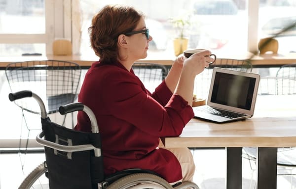 Woman in red robe sitting in wheelchair holding a cup of coffee at a table with a laptop