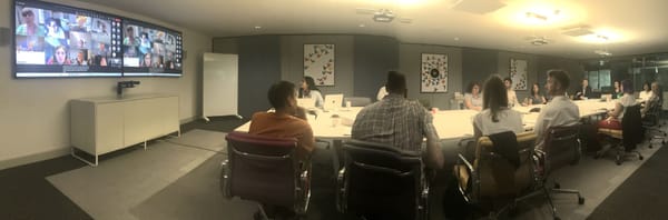 A number of peoiplke sat around a meeting table with a screen of other attendees in smaller pictures.